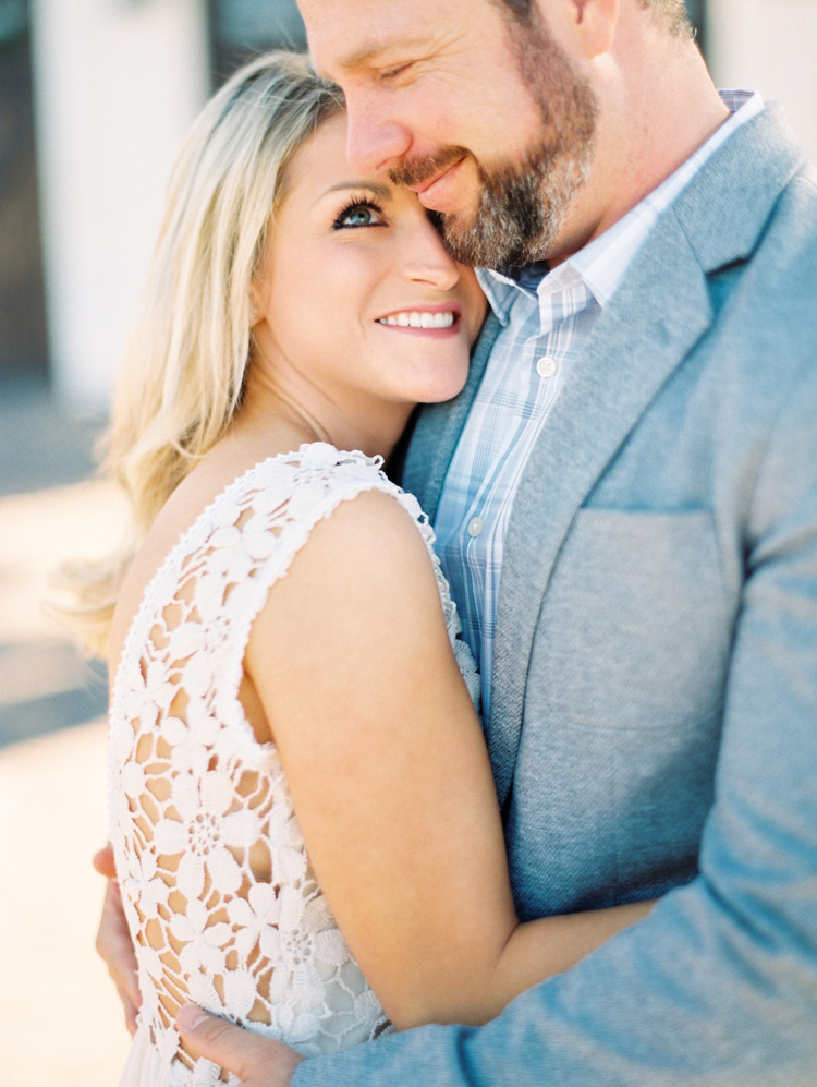 Bride-to-be in a lacy crocheted dress cuddles close to her groom-to-be in a gray sport coat. 