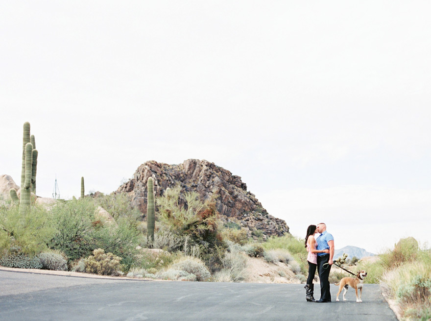 A couple stops for a kiss as they walk their dog near the Arizona desert. Outdoor engagement shoot. 
