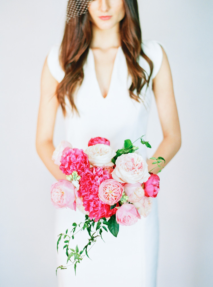 Modern v-neck gown, birdcage veil, and small bouquet of vivid peonies in shades of pink.