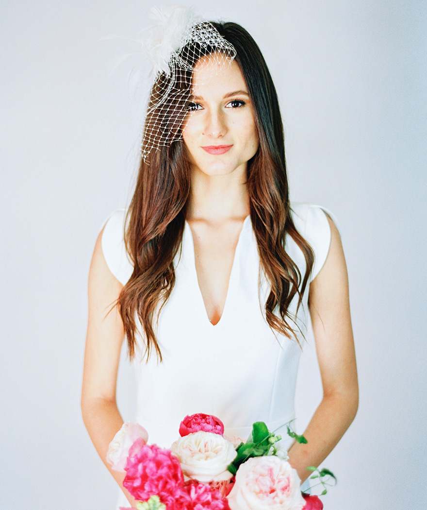 Modern v-neck gown, birdcage veil, and small bouquet of vivid peonies in shades of pink.