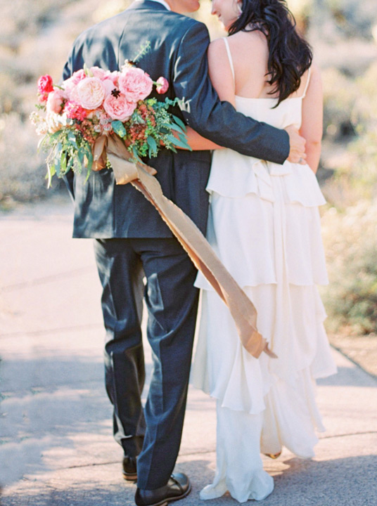 Bride and groom walking through desert landscape at destination wedding. Pink flower bouquet.   