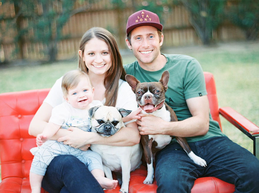 Sweet young couple and baby pose with two dogs on red sofa. Outdoor scenery.