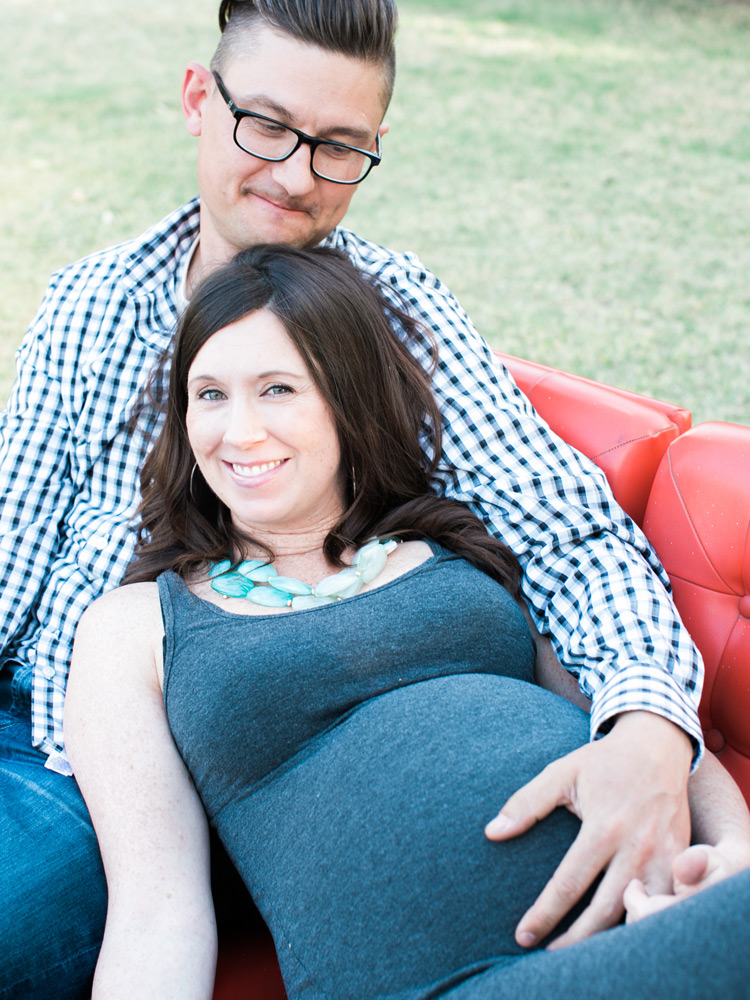 Husband embraces pregnant wife laying on red sofa. Sweet moment outdoors.