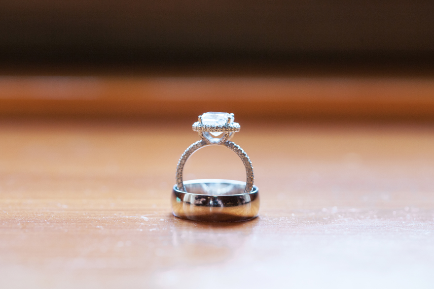 Engagement ring with a halo setting and open gallery sits upright in the groom's wedding band.