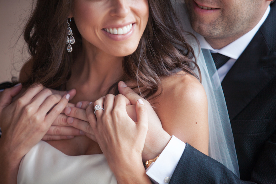 Close-up portrait of the groom embracing his bride from behind. Focus on the engagement ring