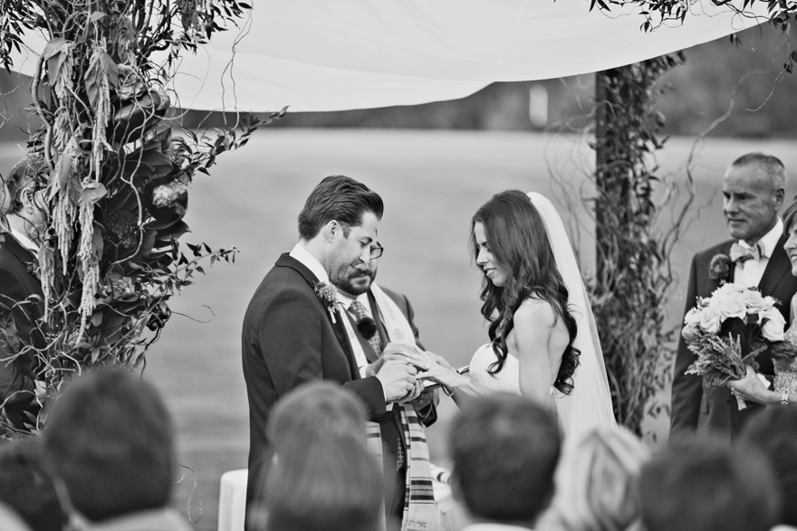Groom slips a ring onto his bride's finger. Black and white photo of an outdoor wedding ceremony