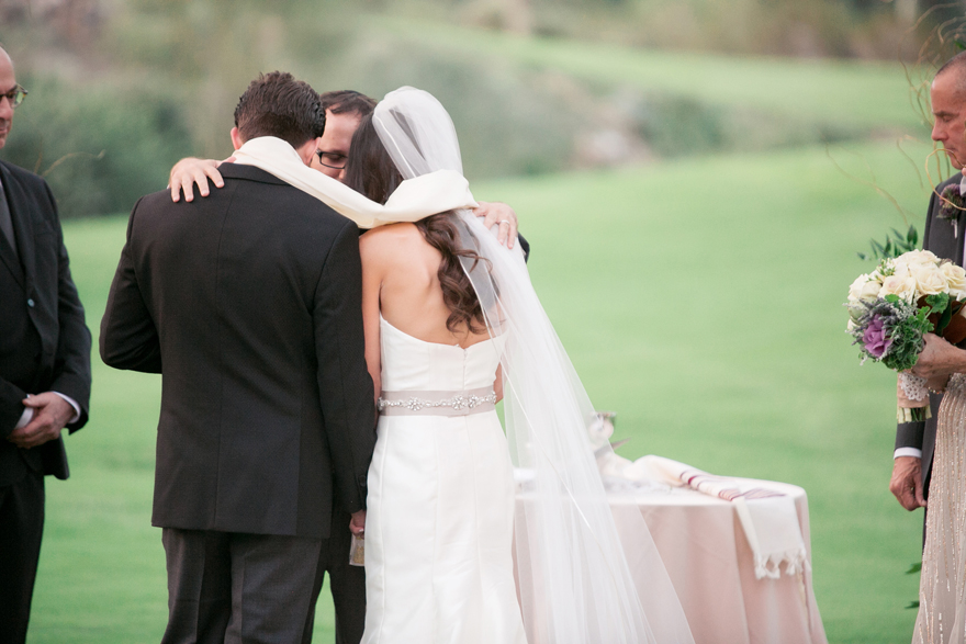 Bride and groom are draped in a prayer shawl as the rabbi recites the blessing. 