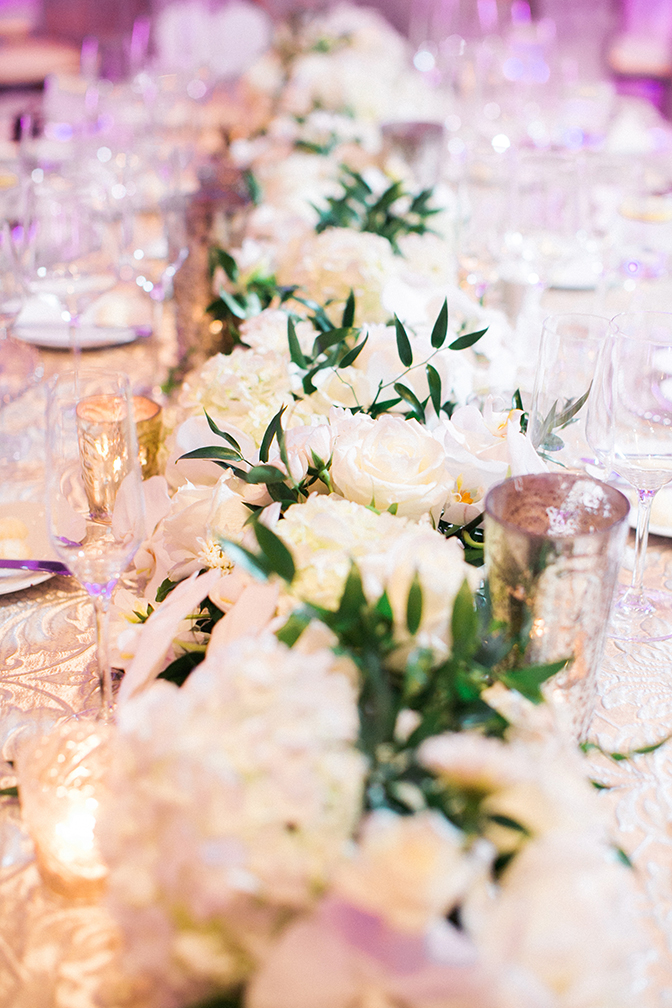 A long runner of white flowers down the center of a table covered in damask. Elegant wedding