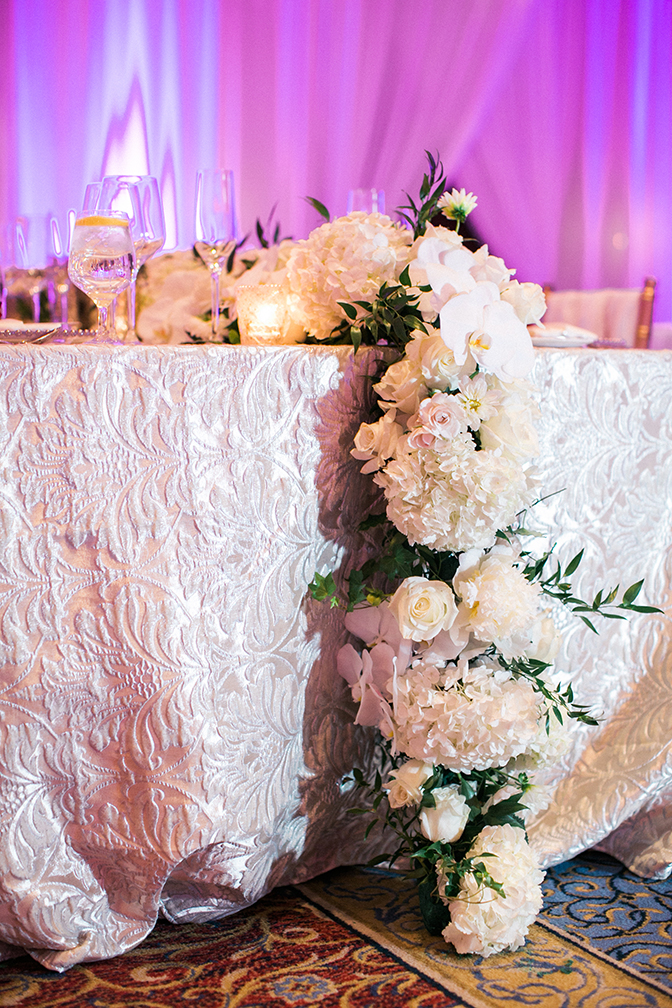 A long runner of white flowers cascades from a table covered in damask. Elegant wedding reception